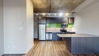 Located on the fourth floor at 10620 116 Street NW in Edmonton, this one-bedroom, one-bathroom unit offers a compact yet efficiently designed layout with an industrial-modern aesthetic. The open-concept living and kitchen area is anchored by an exposed concrete ceiling with track lighting, lending a loft-like character to the space. Wood-look plank flooring runs continuously throughout, adding warmth and visual cohesion.

The kitchen is a standout feature, equipped with dark flat-panel cabinetry accented by vibrant lime-green upper cabinet panels, a light quartz countertop, stainless steel appliances including a range, microwave, dishwasher, and refrigerator, and a peninsula that provides additional counter space and separates the kitchen from the living area.

The bedroom is a well-proportioned private retreat with a window offering views of the surrounding neighbourhood, a baseboard heater, and direct access to a closet fitted with wire shelving and hanging rods. Adjacent to the bedroom, the bathroom features a soaker tub, a floating dark wood vanity with a quartz countertop, a mirrored medicine cabinet, and wood-tile flooring. A hallway connects all areas of the unit, contributing to a practical and logical flow throughout.