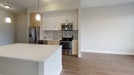 Located at 150 Rivers Edge in Medford, MA, this single-floor residence showcases a clean, modern design with thoughtful finishes throughout. The open-concept layout seamlessly connects the kitchen and living area, creating a bright and functional space. The kitchen is a standout feature, equipped with stainless steel appliances including a refrigerator, electric range, and over-the-range microwave. A large center island with a quartz countertop provides ample workspace and casual seating, while a mosaic tile backsplash adds visual interest behind the range. Cabinetry combines white upper cabinets with warm wood-toned lower units for a contemporary two-tone look, accented by pendant lighting overhead. Hardwood-style flooring flows throughout the main living areas, contributing to a warm, cohesive aesthetic. The living area is generously proportioned, filled with natural light from large windows dressed with horizontal blinds. Bedrooms are finished with plush carpeting and feature walk-in closets with built-in shelving. The bathroom is finished with large-format tile flooring, a floating white vanity with granite countertops, and chrome fixtures, lending a sleek, spa-like quality to the space.