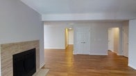 This single-floor space offers an open and functional layout centered around a warm, inviting living area. The flooring throughout is hardwood in a warm honey tone, providing a cohesive and clean aesthetic across the connected spaces. A fireplace with a neutral tile surround serves as a notable focal point along one wall, adding both character and practicality to the living area. The textured popcorn ceiling and crisp white walls create a classic interior backdrop, while recessed lighting ensures adequate illumination throughout. A classic six-panel white door leads to an adjacent area, hinting at the spatial continuity of the floor plan. The open sightlines between rooms contribute to a sense of spaciousness, with a secondary area visible beyond the main living space, suggesting a flexible layout suitable for multiple uses. White baseboards frame the hardwood floors neatly throughout. Overall, the space presents a neutral, move-in-ready environment with practical finishes and a straightforward, well-connected single-level design.