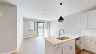 Located at 2120 NW Lowenstein Dr in Lee's Summit, Missouri, this thoughtfully designed single-floor residence offers a clean, contemporary aesthetic throughout. The open-concept layout connects the kitchen and living area seamlessly, creating an inviting, functional space bathed in natural light from large sliding glass doors and windows fitted with vertical blinds. The living area features light wood-tone flooring that extends throughout the entire unit, contributing to a bright and cohesive atmosphere.

The kitchen is well-appointed with crisp white shaker-style cabinetry, quartz countertops, a double-basin undermount sink with a brushed gold pull-down faucet, and a full suite of stainless steel appliances including a refrigerator with ice and water dispenser, electric range, over-the-range microwave, and dishwasher. A stylish conical pendant light accents the kitchen island.

The bedrooms are generously sized, each featuring ceiling fans, natural light from exterior windows, and sliding panel closets with built-in shelving. The bathroom includes a wide vanity with quartz countertop, a large mirror, white cabinetry, and a walk-in shower. Neutral warm-gray wall tones and clean white trim throughout give the space a modern, move-in-ready feel.