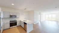 This single-floor residence offers a clean, contemporary design characterized by bright white walls, light-toned flooring, and an open, airy layout. The kitchen serves as a central highlight, featuring an L-shaped configuration with a large peninsula island equipped with a undermount sink and pendant lighting above. White shaker-style cabinetry extends to the ceiling, providing ample storage, while a full suite of stainless steel appliances — including a refrigerator, range, microwave, and dishwasher — delivers a polished, functional finish. Light hardwood-style flooring flows throughout the kitchen and main living areas, which benefit from generous natural light streaming in through large windows with views of surrounding greenery. The living space is open and versatile, connecting seamlessly to the kitchen. The bedrooms are carpeted in a neutral tone, each offering a calm retreat with well-sized windows that invite natural light. The bathroom is finished with a quartz countertop, an undermount sink, a large mirror, and a glass-enclosed shower. A dedicated in-unit laundry room houses a full-size side-by-side washer and dryer. The overall ambiance is bright, modern, and move-in ready.