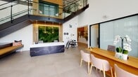 This contemporary two-story space in Durham, North Carolina, makes a striking first impression with its soaring double-height lobby. A standout living green wall serves as the focal point behind a sculptural navy reception desk adorned with delicate metallic detailing. Warm pendant lighting descends from above, complementing the wood-paneled accent walls, while a glass-paneled staircase with sleek black metal railings connects the two floors, adding both visual drama and architectural interest.

The lobby level extends into a well-appointed mailroom featuring geometric patterned accent walls in soft blue, warm wood millwork surrounding built-in mail compartments, and elegant multi-arm brass chandeliers that cast inviting light throughout the space. Large windows allow natural light to filter in, enhancing the open, airy atmosphere.

The upper floor hosts carpeted corridors leading to private office spaces, each thoughtfully furnished. One features a natural wood desk, ergonomic seating, and curated artwork, while another offers a tufted lounge chair beside floor-to-ceiling windows with street-level views. A dining area with a dark round table, wood-toned flooring, and a sculptural pendant light adds a refined social setting. Throughout both floors, a cohesive palette of warm neutrals, earthy tones, and modern finishes creates a polished, welcoming environment.