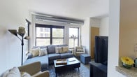 This single-floor space showcases a thoughtfully designed living area that blends modern comfort with industrial-inspired architectural details. Exposed ductwork runs along the ceiling, adding an urban character to the otherwise clean, white-painted surfaces above. Large, black-framed windows dominate one wall, flooding the room with generous natural light and offering views of the surrounding cityscape and greenery beyond.

The living area is well-appointed with a spacious sectional sofa in soft grey, complemented by coordinating accent chairs and decorative throw pillows in neutral and botanical tones. A low-profile dark wood coffee table anchors the seating arrangement atop a patterned area rug, while a large flat-screen television is positioned on a media console opposite the sofa, creating a practical and cohesive entertainment setup.

A wall-mounted air conditioning unit on the left side and a baseboard heater beneath the windows suggest year-round climate control throughout the space. The layout feels open and well-proportioned, with clean sightlines from the main entrance to the living area. Overall, the space offers a contemporary urban atmosphere with practical amenities and ample natural light.