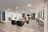 This thoughtfully designed co-working space occupies a single floor and seamlessly blends collaborative and private work environments. The open-plan layout centers around a welcoming lounge area furnished with tufted leather sofas, upholstered armchairs, and a solid wood coffee table atop a textured area rug — ideal for informal meetings or relaxed work sessions. Built-in bookshelves flank a wall-mounted display and a compact kitchenette with cabinetry and a mini-fridge, adding both function and character to the space.

Wide-plank hardwood-style flooring flows throughout, connecting the lounge to a long communal workbench lined with ergonomic seating and pendant lighting — a dedicated zone for focused, side-by-side work. Natural light filters in through a series of black-framed windows that run along one wall, creating a bright and energizing atmosphere.

Private and semi-private work areas include individual focus nooks, each outfitted with a built-in desk, rolling chair, and a pendant light above framed nature-inspired artwork. Glass-walled meeting rooms with black steel frames provide visual transparency while offering acoustic separation, accommodating small group collaboration. The clean white walls, recessed lighting, and contemporary finishes throughout create a cohesive, professional ambiance well-suited for productivity.