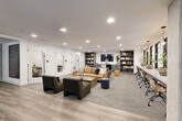 This thoughtfully designed co-working space occupies a single floor and seamlessly blends collaborative and private work environments. The open-plan layout centers around a welcoming lounge area furnished with tufted leather sofas, upholstered armchairs, and a solid wood coffee table atop a textured area rug — ideal for informal meetings or relaxed work sessions. Built-in bookshelves flank a wall-mounted display and a compact kitchenette with cabinetry and a mini-fridge, adding both function and character to the space.

Wide-plank hardwood-style flooring flows throughout, connecting the lounge to a long communal workbench lined with ergonomic seating and pendant lighting — a dedicated zone for focused, side-by-side work. Natural light filters in through a series of black-framed windows that run along one wall, creating a bright and energizing atmosphere.

Private and semi-private work areas include individual focus nooks, each outfitted with a built-in desk, rolling chair, and a pendant light above framed nature-inspired artwork. Glass-walled meeting rooms with black steel frames provide visual transparency while offering acoustic separation, accommodating small group collaboration. The clean white walls, recessed lighting, and contemporary finishes throughout create a cohesive, professional ambiance well-suited for productivity.