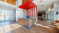 This single-floor community clubhouse offers a generously sized, thoughtfully designed gathering space that blends bold aesthetics with practical amenities. The entrance corridor welcomes visitors with wood-look tile flooring, large glass doors that invite natural light, and an informational display wall, setting an organized and inviting tone from the start.

At the heart of the space is a striking reception desk framed by dramatic red-painted overhead beams — a bold architectural focal point that draws the eye upward and adds a vibrant energy to the interior. Adjacent to the reception area, a comfortable lounge features a mix of leather and upholstered seating arranged around a coffee table, with oversized windows offering views of the surrounding neighborhood and filling the room with abundant natural light.

The game and social area impresses with soaring double-height ceilings, clerestory windows, a full-sized billiards table, and a bar-height counter with seating, all anchored by warm wood-accented ceiling panels. A reclaimed wood feature wall housing a large flat-screen TV adds a rustic contrast to the otherwise modern design.

Completing the space is a functional kitchen equipped with a range, microwave, and ample cabinetry, alongside a beverage station — making this clubhouse a fully self-sufficient hub for community gatherings and everyday use.