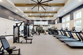 This modern fitness center in Dalton, Georgia, offers a generously sized, single-floor workout facility designed to accommodate a full range of exercise needs. The space is defined by its striking architectural details, including elevated coffered ceilings adorned with rustic exposed wood beams and oversized ceiling fans that maintain comfortable airflow throughout. A bold navy blue and crisp white color scheme lends the space a polished, upscale aesthetic.

The main workout area is well-organized into distinct zones. A comprehensive cardio section lines the perimeter, featuring multiple treadmills, elliptical trainers, and stationary bikes positioned alongside large windows that flood the space with natural light and offer views of the surrounding community. The strength training area is fully equipped with cable machines, a multi-functional rack system, weight benches, and a complete dumbbell rack, providing versatility for users of all fitness levels.

A dedicated functional training room features floor-to-ceiling mirrors, medicine ball racks, kettlebells, and additional specialty equipment, making it ideal for dynamic and group-style workouts. Neutral-toned carpet tiles cover the floors throughout, complementing the overall design. Recessed lighting provides consistent illumination across all areas. The facility is accessible via glass-paneled entry doors and includes a water station for convenience.