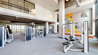 Located at 1000 East Apache Boulevard in Tempe, Arizona, this comprehensive fitness center delivers a well-rounded exercise environment across a generously sized single-floor layout. The interior is defined by striking double-height ceilings, exposed concrete columns, and large floor-to-ceiling windows that flood the space with natural light and offer open views of the surrounding urban landscape. Pendant globe lighting complements the industrial-modern aesthetic throughout.

The main exercise floor is thoughtfully divided into dedicated training zones. A strength and resistance area is equipped with a full lineup of cable machines, multi-use weight benches, and an extensive dumbbell rack, all reflected in wall-to-wall mirrors. A standout feature is the vibrant indoor bouldering wall, accented in yellow and orange, providing a unique vertical challenge within the facility. Crash mats and stabilization equipment round out this area.

Wall-mounted televisions keep members entertained throughout their workout. A staircase connects to an upper-level mezzanine overlooking the main floor.

Accessible via large glass doors, an outdoor functional training courtyard extends the facility's capabilities. Partially shaded by a sail canopy, this open-air space features air bikes, battle ropes, parallette bars, kettlebells, a heavy tire, and a rope climb rig — ideal for high-intensity and cross-training workouts.