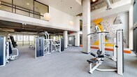 Located at 1000 East Apache Boulevard in Tempe, Arizona, this comprehensive fitness center delivers a well-rounded exercise environment across a generously sized single-floor layout. The interior is defined by striking double-height ceilings, exposed concrete columns, and large floor-to-ceiling windows that flood the space with natural light and offer open views of the surrounding urban landscape. Pendant globe lighting complements the industrial-modern aesthetic throughout.

The main exercise floor is thoughtfully divided into dedicated training zones. A strength and resistance area is equipped with a full lineup of cable machines, multi-use weight benches, and an extensive dumbbell rack, all reflected in wall-to-wall mirrors. A standout feature is the vibrant indoor bouldering wall, accented in yellow and orange, providing a unique vertical challenge within the facility. Crash mats and stabilization equipment round out this area.

Wall-mounted televisions keep members entertained throughout their workout. A staircase connects to an upper-level mezzanine overlooking the main floor.

Accessible via large glass doors, an outdoor functional training courtyard extends the facility's capabilities. Partially shaded by a sail canopy, this open-air space features air bikes, battle ropes, parallette bars, kettlebells, a heavy tire, and a rope climb rig — ideal for high-intensity and cross-training workouts.
