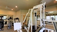 Located at 5555 Tinkey Park in Columbus, Ohio, this fitness center offers a well-equipped, single-floor exercise facility designed to support a comprehensive workout experience. The space is anchored by a prominent white multi-station cable machine, providing users with a wide variety of strength training options. A treadmill is also positioned within the room, catering to cardiovascular exercise needs. Mirrored walls line the space, enhancing the sense of openness while allowing users to monitor their form during workouts. The room features recessed lighting in the ceiling, contributing to a bright and well-lit environment, complemented by a patterned carpet underfoot. French doors with glass panels lead to an adjacent area, adding an architectural detail that creates a sense of flow and connectivity within the facility. The warm-toned walls and clean white equipment give the space a polished and inviting atmosphere. Overall, this fitness center is thoughtfully arranged to maximize functionality while maintaining a comfortable and welcoming environment for users pursuing their health and wellness goals.