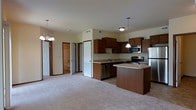 Located at 110 NE 46th Ln in Ankeny, Iowa, this single-floor residence offers a well-designed, open-concept layout that seamlessly connects the kitchen and living areas. The kitchen is a standout feature, equipped with stainless steel appliances including a range, microwave, dishwasher, and refrigerator. Rich, dark wood cabinetry provides ample storage, while a center island with a breakfast bar offers additional workspace and casual dining options. Pendant lighting above the island and a chandelier in the adjoining living area add a warm, inviting ambiance. The living space is finished with neutral carpet that extends throughout, contributing to a cohesive and comfortable feel. The open sightlines from the kitchen into the living area create a sense of spaciousness. A hallway off the main living area leads to additional rooms, including a bathroom visible in the background, featuring tile flooring. A large closet with shelving provides generous storage. The overall design balances functionality with a clean, understated aesthetic, making it a practical and comfortable living space.