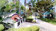This well-appointed residential complex presents a striking combination of contemporary architecture and resort-style outdoor living. The buildings feature a clean white exterior accented by bold teal/blue balconies and railings, creating a visually dynamic façade surrounded by mature trees and verdant landscaping.

The grounds are thoughtfully designed with wide, paved pedestrian walkways that wind through manicured lawns, neatly trimmed hedges, and a rich variety of tropical plantings including towering palm trees, banana plants, and ornamental grasses. The lush greenery creates a sense of privacy and tranquility throughout the community.

A standout feature of the outdoor space is a beautifully tiled circular fountain, adorned with colorful Talavera-style mosaic tilework in deep cobalt blue and terracotta tones, framed by swaying palm fronds. Nearby, a dedicated outdoor BBQ area offers a matching tile-accented wood-fired oven and gas grills, providing an ideal setting for communal gatherings.

Additional amenities include a pool area accessible via stone steps with iron railings, and pathway lighting throughout the grounds for evening use. The overall ambiance is warm, sun-drenched, and resort-like, reflecting a Southern California aesthetic that blends indoor-outdoor living seamlessly.