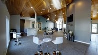 This single-floor space makes an immediate impression with its striking vaulted ceiling clad in warm-toned wood planking, which spans the full length of the open-plan interior and adds a sense of generous vertical volume. Recessed lighting and a central ceiling fan are integrated seamlessly into the wood ceiling, providing both ambiance and practical illumination throughout the day and evening.

The main gathering area is thoughtfully appointed with a mix of seating arrangements, including contemporary bar-height tables paired with sleek white and chrome adjustable stools, as well as a round glass-top table with modern chairs for more intimate settings. Additional lounge seating is arranged along the far end of the space, creating distinct zones for conversation and relaxation.

A built-in bar counter with white bar stools anchors one side of the space, offering a dedicated service or refreshment area. The walls are finished in a sophisticated slate-grey tone that complements the natural warmth of the wood ceiling. Large windows allow natural light to filter in, brightening the interior. Decorative planters with greenery and wall-mounted shelving add finishing touches, while a wall-mounted flat-screen television provides entertainment. The overall ambiance is modern, welcoming, and well-suited for community events or social gatherings.
