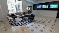 This single-floor lounge space showcases a sophisticated, contemporary design defined by a striking monochromatic color palette of black, white, and gray. The floors are a standout feature, composed of light-toned wood-look tile laid in a herringbone pattern and accented by inlaid metallic square tiles that catch the light and add a glamorous touch throughout the open area. A large seating arrangement anchors the central space, centered on a bold black-and-white geometric circular rug. The seating includes a plush brown sofa adorned with patterned throw pillows, complemented by a sleek black leather chair and a modern white molded accent chair, all surrounding a dark glass coffee table. An oversized metallic arc floor lamp adds a dramatic focal point. Along one wall, a richly textured black decorative panel frames a dual flat-screen television setup, creating a dedicated media feature. Large floor-to-ceiling windows along the opposite wall flood the space with natural light and offer views of the surrounding exterior, enhancing the open, airy atmosphere. The overall ambiance is polished and upscale, balancing bold design elements with functional comfort.