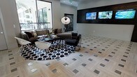 This single-floor lounge space showcases a sophisticated, contemporary design defined by a striking monochromatic color palette of black, white, and gray. The floors are a standout feature, composed of light-toned wood-look tile laid in a herringbone pattern and accented by inlaid metallic square tiles that catch the light and add a glamorous touch throughout the open area. A large seating arrangement anchors the central space, centered on a bold black-and-white geometric circular rug. The seating includes a plush brown sofa adorned with patterned throw pillows, complemented by a sleek black leather chair and a modern white molded accent chair, all surrounding a dark glass coffee table. An oversized metallic arc floor lamp adds a dramatic focal point. Along one wall, a richly textured black decorative panel frames a dual flat-screen television setup, creating a dedicated media feature. Large floor-to-ceiling windows along the opposite wall flood the space with natural light and offer views of the surrounding exterior, enhancing the open, airy atmosphere. The overall ambiance is polished and upscale, balancing bold design elements with functional comfort.