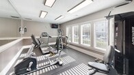 Located at 5715 Castlebridge Rd in Fredericksburg, Virginia, this single-floor fitness center is designed to provide a functional and well-equipped exercise environment. The space features neutral gray walls accented by white trim, a textured ceiling with recessed panel lighting, and durable rubber flooring that offers both safety and practicality throughout the workout area.

The facility is outfitted with a comprehensive selection of cardio and strength equipment, including Matrix-brand treadmills, elliptical trainers, a recumbent bike, and a Bodycraft multi-function cable machine. A wall-mounted flat-screen television is positioned for visibility from the cardio stations, allowing users to stay entertained during workouts. Large, white-framed windows fitted with horizontal blinds line one wall, allowing ample natural light to fill the space and providing views to the outdoors. A full-length wall mirror on the opposite side visually expands the room and supports proper form during exercise. The space also includes a sanitation station for equipment maintenance. The overall ambiance is clean, bright, and purposefully arranged, making it a practical and welcoming fitness resource for the surrounding community.