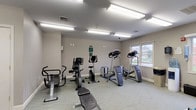 The fitness center at 143 Shore Rd in Peabody, Massachusetts is a well-equipped, single-floor exercise facility designed to support a comprehensive workout routine. The room is finished with neutral gray walls and durable patterned carpet flooring, creating a clean and functional environment throughout.

The cardio section is generously stocked, featuring multiple treadmills, stair climbers, elliptical machines, and stationary bikes — including both upright and recumbent models — providing a wide variety of low-impact and high-intensity cardiovascular options. A multi-station strength machine and a dedicated free weights area with a dumbbell rack and kettlebells round out the strength training offerings, while a weight bench provides additional versatility.

Bright fluorescent lighting illuminates the space evenly, and multiple windows along the exterior walls allow natural light to filter in, offering views of the surrounding outdoor area. A wall-mounted television is positioned for easy viewing during workouts. Practical amenities include a water cooler, paper towel dispensers, and a waste bin, ensuring convenience during use.

The facility is clearly marked with safety and emergency signage, including exit indicators and fire safety equipment. Overall, the fitness center is thoughtfully arranged to maximize usable floor space while offering a broad spectrum of equipment for residents seeking a complete on-site workout experience.