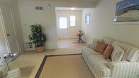 This 2-floor home offers a warm and functional layout designed for everyday comfort. The living room serves as the heart of the main level, featuring wall-to-wall carpeting in a soft neutral tone that adds warmth underfoot. A large area rug with a classic border detail anchors the seating area, complemented by a striped sofa dressed with accent pillows. A lush potted plant in the corner introduces a natural element, while a baseboard heater and ceiling-mounted HVAC vent ensure year-round climate comfort. The living room opens seamlessly into a defined entryway, creating an inviting transition space. The front entry features a paneled door flanked by windows that allow natural light to filter in, brightening the adjoining area. A small decorative accent table with a delicate floral arrangement adds a charming touch near the entrance. The overall color palette throughout is soft and neutral, with light beige walls and carpeting that create a cohesive, calming ambiance. The open sightlines between the living room and entryway enhance the sense of space, making the home feel both connected and airy across its two levels.