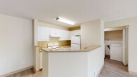 This one-bedroom, one-bathroom unit in Beaverton, Oregon offers a well-organized single-floor layout designed for comfortable everyday living. The open-concept kitchen and living area serves as the heart of the home, featuring a galley-style kitchen equipped with white cabinetry, laminate countertops, a full suite of white appliances including a refrigerator and electric range, and a wraparound peninsula that provides additional prep and dining space. Wood-style flooring runs throughout, lending a cohesive and clean aesthetic to the interior. The living room is a standout area, highlighted by a tile-surround fireplace and large sliding glass doors flanked by a window, filling the space with natural light and offering a view of the outdoors. The bedroom is a comfortable size, featuring its own window with vertical blinds and direct access to a closet. The full bathroom includes a single-sink vanity with white cabinetry, a wide mirror, and a tub/shower combination. A dedicated in-unit laundry area accommodates a full-size washer and dryer side by side, adding a practical convenience. Warm neutral tones on the walls carry throughout the unit, creating a calm and inviting atmosphere.
