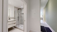 This thoughtfully designed single-floor unit offers an efficient yet stylish layout that maximizes every square foot. The open-concept kitchen and living area serves as the heart of the space, where a well-appointed kitchen features shaker-style cabinetry with glass-front upper panels, a marble-look backsplash, stainless steel appliances, and light hardwood flooring. Exposed ductwork overhead adds an industrial touch that contrasts attractively with the otherwise refined finishes. A compact dining area with a round wood table transitions naturally into the living room, which is distinguished by a soft sage-green accent wall, large windows that invite natural light and outdoor views, and warm, layered décor. Both bathrooms maintain the unit's modern aesthetic, featuring frameless glass-enclosed showers, large-format tile surrounds, wall-mounted floating vanities with illuminated mirrors, and light hardwood-style flooring. A dedicated walk-in closet provides well-organized built-in storage with hanging rails, shelving, and integrated drawers. Throughout the unit, a neutral palette of whites and soft greens creates a cohesive, calming ambiance.