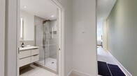 This thoughtfully designed single-floor unit offers an efficient yet stylish layout that maximizes every square foot. The open-concept kitchen and living area serves as the heart of the space, where a well-appointed kitchen features shaker-style cabinetry with glass-front upper panels, a marble-look backsplash, stainless steel appliances, and light hardwood flooring. Exposed ductwork overhead adds an industrial touch that contrasts attractively with the otherwise refined finishes. A compact dining area with a round wood table transitions naturally into the living room, which is distinguished by a soft sage-green accent wall, large windows that invite natural light and outdoor views, and warm, layered décor. Both bathrooms maintain the unit's modern aesthetic, featuring frameless glass-enclosed showers, large-format tile surrounds, wall-mounted floating vanities with illuminated mirrors, and light hardwood-style flooring. A dedicated walk-in closet provides well-organized built-in storage with hanging rails, shelving, and integrated drawers. Throughout the unit, a neutral palette of whites and soft greens creates a cohesive, calming ambiance.