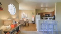 This single-floor residence showcases an open-concept design that effortlessly connects the main living spaces, creating a cohesive and functional layout. The kitchen is equipped with warm medium-toned cabinetry, a full suite of appliances including a refrigerator, microwave, and range, and features light granite countertops that extend into a raised breakfast bar with modern bar seating — ideal for casual dining or entertaining. Track lighting overhead ensures the kitchen area is well-lit and practical. Flowing directly from the kitchen, the dining area is defined by warm hardwood-style flooring and anchored by a statement chandelier that draws the eye upward. A bold decorative sunburst wall piece adds an artistic focal point to the space. Chevron-patterned dining chairs lend a touch of contemporary style, while neutral wall tones keep the atmosphere open and inviting. The transition into the adjacent living area is smooth, with plush carpeting providing a comfortable contrast to the hard flooring of the dining and kitchen zones. Natural light filters in through a prominently arched window, brightening the space. The overall ambiance is warm, modern, and well-balanced, with thoughtful design details throughout.