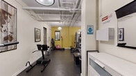 Located at 1851 Ivar Ave in the heart of Hollywood, this single-floor exercise room offers a comprehensive fitness facility designed to support a wide range of workout routines. The space features durable rubber flooring throughout, providing excellent traction and cushioning for both cardio and strength training activities.

The room is appointed with a diverse selection of equipment, including treadmills, a stationary bike, and a multi-station cable machine for versatile strength training. Additional equipment includes a power tower, a hyperextension bench, a flat bench, barbells with weight plates, dumbbells, medicine balls, and a speed bag — catering to users of all fitness levels. A wall-mounted television adds convenience for entertainment during workouts.

Full-length mirrors line the walls, amplifying the sense of space and allowing users to monitor their form. The walls are painted in a warm yellow accent tone complemented by white, giving the room an energetic yet clean ambiance. Pendant lighting fixtures provide bright, even illumination throughout. Exposed ceiling pipes lend an industrial character to the space. A decorative world map artwork and framed pieces add a finishing touch to the room's aesthetic, making this fitness center both functional and visually engaging.