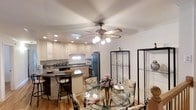 This single-floor residence showcases an open-concept layout that seamlessly connects the kitchen and dining area, creating a cohesive and functional living space. The kitchen is appointed with cream-toned cabinetry featuring raised-panel doors, dark granite countertops, and recessed lighting that provides ample illumination. A curved breakfast bar with dark granite extends from the kitchen island, accompanied by tall bar stools, offering a casual seating option. Stainless and black appliances, including a full-size refrigerator and built-in microwave, complement the kitchen's practical design. Light hardwood flooring flows throughout the space, adding warmth and continuity. The adjacent dining area accommodates a glass-top dining table with ornate metal legs, surrounded by upholstered floral chairs that lend a traditional charm. A decorative ceiling fan with a light fixture serves as a central focal point overhead, while two black metal display shelving units frame the dining area, providing stylish storage. Crown molding along the ceiling adds a refined architectural detail, enhancing the overall elegance of the space.