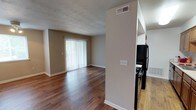 This single-floor space offers a practical and functional layout with a clear separation between the living area and kitchen. The living room is a standout feature, showcasing rich, dark hardwood-style flooring that extends throughout the space, creating a warm and cohesive aesthetic. A large sliding glass door fitted with vertical blinds serves as the focal point of the living area, allowing natural light to fill the room while providing access to an outdoor area.

The textured ceiling adds a subtle design element overhead, while neutral-toned walls in a warm beige palette keep the space feeling bright and versatile. A ceiling vent indicates a central HVAC system for year-round climate control.

A partial wall divides the living area from the kitchen, offering a degree of separation while maintaining an open feel. The kitchen features lighter hardwood-style flooring, contrasting with the darker tones of the living room. Black appliances, including a refrigerator and stove, are visible, providing a sleek, functional cooking space. The overall layout is efficient and well-suited for everyday living.