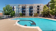 Located at 2550 Waterview Dr in Glenview, Illinois, this thoughtfully designed outdoor amenity space serves as a vibrant communal hub surrounded by a modern multi-story residential building. The courtyard is anchored by a generously sized swimming pool, complemented by ample deck space ideal for lounging and recreation. Sturdy pergola structures with clean-lined, contemporary frames provide shaded gathering areas throughout, casting pleasant dappled light across the open walkways below. A dedicated grilling area equipped with multiple gas grills and outdoor dining furniture offers a convenient space for outdoor entertaining. Raised stone planter walls constructed from natural, multi-toned stonework define the various zones within the courtyard, adding a distinctive and durable architectural element to the landscape. Lush, mature plantings — including ornamental grasses, flowering shrubs, and deciduous trees — are woven throughout, softening the hardscape and providing greenery and seasonal color. Wide concrete pathways connect each zone seamlessly, ensuring easy circulation throughout the space. The building's contemporary facade, featuring blue and gray siding, large windows, and private balconies, frames the courtyard on multiple sides, creating an enclosed yet open-air atmosphere. The overall ambiance is clean, modern, and inviting, offering residents a well-appointed outdoor retreat.