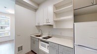 This efficiently designed single-floor studio offers a well-considered layout that maximizes every square foot of its compact footprint. The open main living area features light wood-look flooring and a soft two-tone color palette, with crisp white walls complemented by a muted blue-grey accent wall that adds a touch of modern character. A large window allows natural light to flow into the living space, creating a bright and airy atmosphere.

The kitchen is thoughtfully appointed and makes excellent use of its dedicated space. Two-tone cabinetry pairs white upper cabinets with grey lower cabinets, all fitted with sleek silver bar hardware. Light quartz countertops provide a clean, contemporary work surface. The kitchen includes a stainless steel sink, a built-in cooktop, an under-counter microwave, a refrigerator, and a range hood, along with open upper shelving for additional storage.

The bathroom continues the clean, minimal aesthetic of the space, featuring light wood-look flooring, a white vanity with an integrated sink, and a wall-mounted grab bar, suggesting accessibility-conscious design. White walls keep the space feeling open and hygienic.

Throughout the studio, the design prioritizes functionality and simplicity, making it a practical and comfortable living environment.
