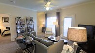 This single-floor space offers a thoughtfully designed interior that balances comfort and style. The living room serves as the centerpiece, showcasing crown molding along the ceiling and a classic ceiling fan with integrated lighting that adds both function and charm. Large windows dressed with elegant gray curtains and blinds flood the space with natural light, creating a warm and inviting atmosphere. A glass-paneled door provides additional brightness and potential outdoor access. The living area is generously proportioned, accommodating a plush sectional sofa in a neutral gray tone, complemented by a zebra-print area rug and a sleek oval glass coffee table. A cozy armchair is positioned near the windows, creating a relaxing reading nook. A tall leaning bookshelf along one wall provides display and storage space, adding a decorative and functional element to the room. Soft, neutral wall tones throughout enhance the sense of openness, while carefully placed floor lamps and table lamps contribute to a layered lighting scheme. The overall ambiance is polished and contemporary, with cohesive furnishings and accessories that reflect a refined, modern aesthetic throughout the space.