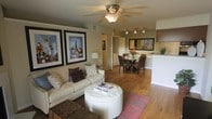 This single-story residence showcases an open-concept floor plan that seamlessly connects the living, dining, and kitchen areas into one cohesive and functional space. The living area is anchored by a plush, neutral-toned sofa accented with decorative pillows and paired with a round tufted ottoman that doubles as a coffee table. A bold area rug adds a pop of color beneath, while two large framed art pieces provide a striking focal point on the main wall. A ceiling fan with an integrated light fixture keeps the space comfortable and well-lit. Warm hardwood-style flooring flows throughout, lending continuity and a welcoming feel to the entire layout. The dining area features a glass-top table surrounded by woven rattan chairs, creating an inviting spot for meals. A decorative mirror and fresh floral arrangement enhance the ambiance. Adjacent to the dining space, the kitchen offers light cabinetry, a breakfast bar, and track lighting overhead, making it both practical and stylish. The overall ambiance is warm, contemporary, and well-appointed.