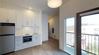 This thoughtfully designed single-floor space showcases a clean, modern aesthetic throughout. The open layout is anchored by a compact yet fully equipped kitchenette featuring crisp white cabinetry with sleek hardware, a built-in cooktop, undermount sink, and a stainless steel microwave oven. The cabinetry extends both above and below the counter, offering ample storage in a minimalist footprint. Light wood-tone flooring runs throughout the space, lending warmth and continuity to the interior. A pendant light fixture overhead provides focused illumination, complementing the recessed lighting built into the ceiling. A hallway leads further into the unit, hinting at additional rooms beyond, with a wall-mounted panel serving as a functional utility feature. One of the standout elements of this space is a glass-paneled door that opens to a private balcony, allowing natural light to pour in and providing a direct connection to the outdoors. The bright, neutral color palette of white walls and ceilings keeps the interior feeling airy and open. Overall, the space presents a contemporary and efficient living environment with clean lines and practical design choices throughout.