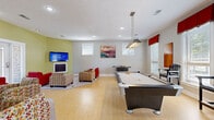 Located at 1000 Worthington Ln in Spring Hill, Tennessee, this single-floor community living and recreation room offers a well-rounded entertainment experience within a generously open layout. The space is finished with light hardwood-style flooring throughout, contributing to a bright and welcoming atmosphere complemented by recessed lighting and a modern pendant light above the pool table area.

The room is distinctly divided into two functional zones. The lounge area features bold red upholstered seating and vibrant patterned armchairs arranged around a area rug, anchored by a wall-mounted flat-screen TV above a fireplace with classic cabinet-style doors. French doors along one wall provide a view to an outdoor area, flooding the space with natural light. Colorful abstract artwork adorns the walls, adding a lively aesthetic throughout.

The game zone is equally well-appointed, housing a full-size pool table with an industrial-style overhead light, a foosball table, and additional bar-height seating along the windowed wall. Multiple large windows dressed with red valances allow ample natural light to fill the area. The space also includes clearly marked exits and access doors, reflecting its community-use function. Overall, the room delivers a dynamic, social environment ideal for recreation and gathering.
