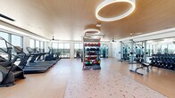 Located at 3238 Hidden Forest Ct in Marietta, GA, this well-appointed fitness center offers a generous single-floor layout designed to accommodate a comprehensive range of workout routines. The space is bathed in natural light courtesy of expansive floor-to-ceiling windows that line multiple walls, providing open views of the surrounding landscape and creating an energizing atmosphere for exercise.

The interior features light hardwood-style flooring throughout, complemented by a warm wood-paneled ceiling adorned with large circular pendant light fixtures that provide ample, evenly distributed illumination. Ceiling fans are strategically placed to ensure proper air circulation across the entire floor.

The cardio zone is well-equipped with multiple treadmills, stair climbers, and stationary bikes with integrated display screens, all positioned to take advantage of the panoramic window views. A dedicated functional training area occupies the center of the space, featuring a multi-use rack stocked with kettlebells, medicine balls, resistance bands, and stability balls atop a patterned exercise mat. The strength training section houses a cable machine system, a multi-station cable cross, a full dumbbell rack, and various weight benches, all arranged along mirrored walls to maximize usable space. The layout is thoughtfully organized to allow fluid movement between workout zones.