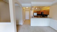 This single-floor residence offers a thoughtfully designed open-concept layout that connects the living area to the kitchen seamlessly. The kitchen is a functional highlight of the space, featuring warm wood-toned cabinetry, dark granite countertops with a complementary tile backsplash, and stainless steel appliances including a refrigerator and microwave. A white peninsula with wainscoting detail provides additional counter space and serves as a natural divider between the kitchen and the carpeted living area. Track lighting illuminates the kitchen, while recessed ceiling details and crown molding add an architectural touch throughout. An arched entryway creates an elegant transition between rooms, enhancing the open feel of the floor plan. The entryway hallway is finished with light-toned tile flooring and leads to interior doors, offering a clean and practical entry point into the home. Neutral wall tones and soft carpeting in the living space contribute to a warm and inviting ambiance throughout the residence.