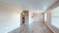 This single-story unit offers a practical and well-organized layout designed to maximize usable space across two bedrooms and one full bathroom. The living area serves as the central hub of the unit, featuring wood-style plank flooring in a warm neutral tone, textured walls painted in soft beige-gray, and a ceiling-mounted light fixture. A built-in niche along one wall adds a subtle architectural detail, while an open passageway connects directly to the kitchen, promoting a natural flow between the living and cooking spaces.

The kitchen is equipped with black appliances, including a full-size refrigerator and dishwasher, complemented by white cabinetry and laminate countertops. Wood-style flooring continues throughout this space, maintaining visual consistency.

Both bedrooms feature plush carpet flooring and neutral gray walls, each receiving natural light through windows fitted with horizontal blinds. The rooms offer a quiet, comfortable atmosphere with ample wall space for furnishings.

The bathroom includes a full-size soaking tub with a tiled surround, a toilet, and an overhead storage cabinet, all finished with the same wood-style flooring found elsewhere in the unit. The overall design emphasizes clean lines, neutral finishes, and a cohesive aesthetic throughout.