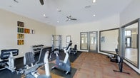 This fitness center offers a comprehensive workout environment designed to accommodate a variety of exercise routines. The space is outfitted with a selection of cardio equipment, including stationary bikes and other aerobic machines, positioned on protective rubber flooring mats that contrast with the surrounding carpet. A multi-functional cable machine and weight rack stocked with a full range of dumbbells provide ample strength training options. The walls are finished in a neutral, light tone that contributes to a clean and open atmosphere. Recessed lighting is installed throughout the ceiling, supplemented by two ceiling fans that promote air circulation during workouts. Natural light filters into the space through a set of glass-paneled double doors, which also provide direct access to the outdoors. Framed informational displays are mounted on the walls, offering guidance and reference material for users. The overall layout is thoughtfully organized, ensuring clear pathways between equipment and an uncluttered workout environment. The combination of natural light, ventilation, and varied equipment makes this fitness center a functional and inviting space for daily exercise.