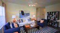 This single-floor space showcases a thoughtfully curated living area that blends contemporary style with vibrant energy. The living room serves as the focal point, anchored by a crisp white sofa adorned with a mix of navy blue and geometric-patterned throw pillows. A pair of deep navy armchairs with bold orange accent pillows frame the seating arrangement, creating a cohesive and striking color palette throughout. A rich walnut-toned coffee table sits at the center, complemented by a bold navy and white hexagonal area rug that adds visual depth and texture to the floor. Vibrant orange table lamps on either side of the room provide warm ambient lighting, while a ceiling fan with an integrated light fixture ensures comfortable airflow. A large-scale abstract canvas painting in blue, black, and orange tones dominates one wall, serving as a dramatic artistic statement that ties the room's color scheme together seamlessly. The walls are finished in a soft, neutral tone that allows the furnishings and décor to take center stage. A hallway visible in the background hints at additional rooms beyond, while the overall layout feels open and well-proportioned, offering a stylish and comfortable environment ideal for both relaxing and entertaining.