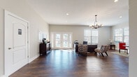 This well-appointed single-floor space is designed to balance professional functionality with an inviting, comfortable atmosphere. The main reception and office area serves as the focal point of the layout, anchored by a large dark executive desk accompanied by two stylishly patterned accent chairs. A decorative wrought-iron chandelier with frosted globe shades hangs overhead, complementing the recessed lighting that illuminates the generous open floor plan. Rich, dark hardwood floors run throughout, providing a warm and polished foundation. French doors at the front of the office area flood the space with natural light while offering views to the outdoors, and additional windows fitted with white horizontal blinds reinforce the bright, airy feel. A dark wood credenza along one wall provides practical storage and a surface for a coffee station, adding a welcoming touch for visitors. Adjacent to the main office, a dedicated meeting or dining area features multiple large windows on two walls, maximizing daylight from multiple angles. This room accommodates a round wood dining table paired with bold red upholstered chairs atop a patterned area rug, creating a vibrant and functional gathering space. Clean gray walls, crisp white trim, and consistent hardwood flooring tie the two areas together cohesively, resulting in a professional yet approachable environment.
