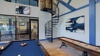 This impressive amenity space combines recreation and fitness in a thoughtfully designed environment. The game room commands attention with its soaring double-height ceilings and a striking wrought-iron spiral staircase that serves as a dramatic architectural centerpiece. Large windows flood the space with natural light, accentuating the warm wood-look flooring and crisp white walls accented with bold blue trim. A billiards table with a rich blue felt surface anchors the room, complemented by a classic shuffleboard table with an industrial-style base, making it an ideal setting for social gatherings. Contemporary abstract artwork in deep navy tones adorns the walls, tying together the space's cohesive color palette. An adjacent fitness center, visible through sleek glass partition walls and doors, offers a well-equipped workout area featuring a dumbbell rack and cardio and strength training equipment. The open, connected layout between the game room and fitness center creates a seamless flow, while the overall design balances an energetic, social atmosphere with functional amenities. The combination of high-end finishes, bold design choices, and versatile recreational offerings makes this a standout communal destination.