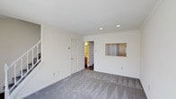 This two-floor residence offers a clean, straightforward layout designed for comfortable everyday living. The interior is characterized by crisp white walls and smooth ceilings that lend a bright, airy feel throughout. Recessed lighting is integrated into the ceiling, providing even, practical illumination across the main living areas. The floors are covered in plush gray carpet with a subtle geometric pattern, adding texture and warmth underfoot. A pass-through window cutout in one wall is a notable architectural detail, offering a convenient visual and functional connection between adjoining spaces. Doors lead to additional rooms, including what appears to be a bathroom or utility area with warm-toned wood flooring visible beyond the threshold. A staircase is accessible from the main living area, connecting the two floors of the home. The overall ambiance is neutral and versatile, providing a blank canvas well-suited to a variety of interior styles. The space is thoughtfully arranged to maximize flow and usability, with multiple access points and thoughtful built-in details that reflect a practical residential design.