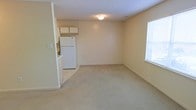 Located at 4016 Copperfield Dr in New Bern, North Carolina, this single-floor residence offers a clean, functional layout designed for comfortable everyday living. The main living area is finished with soft, wall-to-wall carpeting that lends a warm and quiet atmosphere to the space. Neutral cream-toned walls throughout create a bright, open feel that is easy to personalize. A window along the exterior wall allows natural light to filter into the living space, enhancing the airy ambiance. The kitchen is conveniently positioned adjacent to the living area, separated by a partial wall that maintains an open and connected feel between the two spaces. The kitchen features white cabinetry mounted above the counters, providing ample storage, and is equipped with a full-size refrigerator. The light-colored flooring in the kitchen contrasts gently with the carpeted living space, helping to define each functional zone. The textured ceiling throughout adds a classic residential touch. Overall, the space presents a straightforward and well-maintained interior that offers a comfortable, move-in ready environment in a residential New Bern neighborhood.