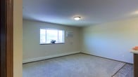 This single-floor space presents a straightforward, functional layout centered around a generously sized room finished with neutral-toned wall-to-wall carpeting. The room is kept bright by a multi-pane window positioned along one wall, allowing natural light to filter in while offering a view of the surrounding outdoor environment. A wall-mounted air conditioning unit is installed adjacent to the window, providing a dedicated climate control solution for warmer months. Along the base of the walls, a baseboard heating system ensures consistent warmth during cooler seasons, offering a dual-climate setup well-suited for year-round comfort. The ceiling is smooth and painted in a light neutral tone, complementing the pale walls throughout. A flush-mount ceiling light fixture provides overhead illumination, keeping the space evenly lit. Dark wood-toned baseboards run along the perimeter, adding a subtle contrast to the light-colored walls and carpet. The overall ambiance is clean, simple, and move-in ready, with a neutral palette that provides a versatile backdrop suitable for a variety of uses and furnishing styles.