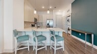 Located at 300 Central Ave in the heart of Sarasota, FL, this single-floor residence combines modern design with coastal-inspired aesthetics. The open-concept kitchen serves as a central hub, featuring light wood cabinetry, white quartz countertops, a subway tile backsplash, stainless steel appliances including a French door refrigerator, and a large island with seating for three. Pendant lighting illuminates the workspace, while wood-look flooring extends seamlessly into the living area. The living room is bright and inviting, benefiting from large windows with plantation shutters that allow natural light to filter in, complemented by patterned drapery and a ceiling fan overhead.

Two well-appointed bedrooms offer distinct personalities: one features bold teal accent walls and decorative lighting, while the other embraces a botanical theme with lush printed curtains and natural light. Both rooms include plush carpeting. A spacious walk-in closet with wire shelving systems provides ample storage. The bathroom is cleanly finished with large-format tile flooring, a soaking tub with shower, and a light wood vanity with white countertops. An in-unit stacked washer and dryer adds everyday convenience.