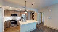 This single-floor residence showcases a thoughtfully designed open layout anchored by a well-appointed kitchen. The kitchen features warm wood-toned cabinetry, a white quartz countertop island with an undermount sink and flexible gooseneck faucet, and a full suite of stainless steel appliances including a French door refrigerator, range, and built-in microwave. Two pendant lights with metallic finishes hang above the island, providing focused task lighting that complements the recessed ceiling lights throughout. A white subway tile backsplash adds a clean, classic detail to the cooking area. Conveniently integrated into the kitchen zone is a stacked washer and dryer unit, offering practical in-unit laundry access.

The bedrooms are finished with durable hardwood-style flooring and neutral white walls, creating bright, flexible living spaces. One bedroom includes a ceiling fan with an integrated light fixture, while another benefits from a large window with outdoor views, allowing natural light to fill the room.

The bathroom is finished in a modern style, featuring light tile flooring, a tub and shower combination clad in white subway tile, a wood-grain vanity with a white countertop, an illuminated LED mirror, and a wall-mounted storage cabinet. Brushed nickel fixtures provide cohesive, contemporary detailing throughout.