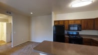 This single-floor residential space is designed with an open-concept layout that seamlessly connects the kitchen and living area, creating a functional and cohesive environment. The kitchen is equipped with rich, warm-toned wood cabinetry that provides ample storage, complemented by sleek black appliances including a refrigerator, range, and over-the-range microwave. A peninsula-style breakfast bar with a durable stone-finish countertop extends into the living space, offering additional workspace and casual dining options. Recessed lighting and a flush-mount ceiling fixture provide even illumination throughout the area, contributing to a well-lit interior. The living area features neutral-toned carpeting that adds warmth and comfort underfoot, while the smooth, light-colored walls create a clean and versatile backdrop. The open floor plan allows for flexible furniture arrangement and encourages a sense of spaciousness throughout. The overall ambiance is practical and inviting, with a neutral color palette that lends itself to a variety of décor styles, making it a comfortable and adaptable living environment.