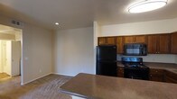 This single-floor residential space is designed with an open-concept layout that seamlessly connects the kitchen and living area, creating a functional and cohesive environment. The kitchen is equipped with rich, warm-toned wood cabinetry that provides ample storage, complemented by sleek black appliances including a refrigerator, range, and over-the-range microwave. A peninsula-style breakfast bar with a durable stone-finish countertop extends into the living space, offering additional workspace and casual dining options. Recessed lighting and a flush-mount ceiling fixture provide even illumination throughout the area, contributing to a well-lit interior. The living area features neutral-toned carpeting that adds warmth and comfort underfoot, while the smooth, light-colored walls create a clean and versatile backdrop. The open floor plan allows for flexible furniture arrangement and encourages a sense of spaciousness throughout. The overall ambiance is practical and inviting, with a neutral color palette that lends itself to a variety of décor styles, making it a comfortable and adaptable living environment.