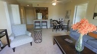 This single-floor residence offers a seamlessly connected open-concept living space that integrates the living room, dining area, and kitchen into one cohesive environment. The living area is finished with soft, plush carpeting in warm neutral tones, creating a welcoming and comfortable atmosphere. A ceiling fan overhead provides both function and a casual elegance to the space.

The kitchen is positioned toward the back of the open layout and features stainless steel appliances, warm wood-toned cabinetry, and a raised breakfast bar with seating for two. The bar stools add a casual dining option, while a dedicated dining area nearby accommodates a full table and chair set, ideal for more formal meals.

Crown molding adds a refined architectural detail along the ceiling, enhancing the overall finish of the space. Recessed lighting in the kitchen, combined with natural light filtering through the windows, keeps the interior bright and inviting. The entryway is visible from the living area, contributing to the open and airy feel of the floor plan. The overall ambiance is cozy yet functional, with a layout that promotes easy movement and everyday living.